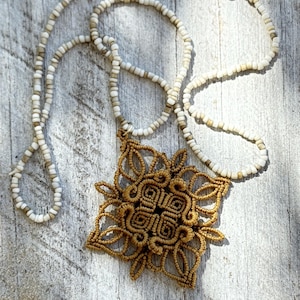Pode incluir: Um colar de macramé feito à mão com um pingente detalhado marrom dourado em um design floral. O colar apresenta uma corrente de contas brancas e um fecho prateado. O colar é exibido em uma superfície de madeira desgastada.