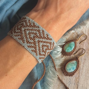 May include: A macrame bracelet in light blue and brown, featuring a geometric design, worn on a wrist. Matching earrings with turquoise stones and brown macrame settings are also visible. The jewellery is displayed on a blue fabric and wooden surface.