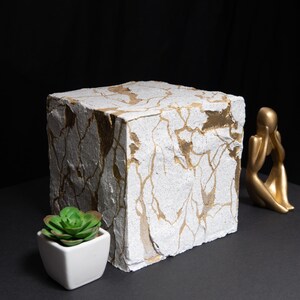 Op de afbeelding: Een groot, wit, kubusvormig decoratief object met goudkleurige, aderachtige patronen. Een kleine, groene vetplant in een witte vierkante pot en een goudkleurig beeldje zijn ook aanwezig. De achtergrond is zwart.