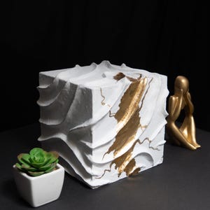 Peut inclure: Un cube blanc texturé avec des accents dorés et un design sculpté. Un petit succulent dans un pot carré blanc et une figurine dorée sont également présents, le tout sur fond sombre.