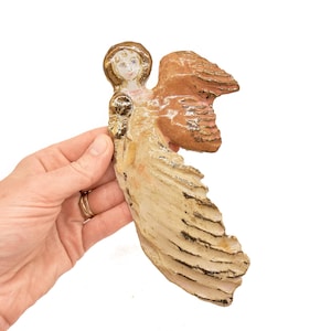 May include: A ceramic angel figurine featuring a glossy cream-colored gown with gold-painted accents, a textured brown wing, and a halo. The figure has a painted face with defined features and a smooth, glazed finish throughout the entire surface.