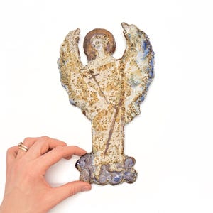 May include: A ceramic angel figurine with outstretched wings, a halo, and a staff. The angel is painted in shades of beige, brown, and blue. The wings have a textured, speckled appearance. The base of the figurine is a cluster of blue and purple shapes.