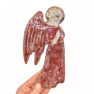 Puede incluir: Una figura de ángel de cerámica con un esmalte rojo brillante y un halo dorado. La figura presenta alas texturizadas y una túnica larga y fluida, con un rostro blanco y rasgos faciales sutiles bajo el halo circular de color dorado.