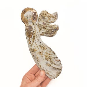 Pode incluir: Uma figura de anjo em cerâmica plana com uma superfície texturizada em tom branco sujo, salpicada com detalhes em castanho e dourado. A figura apresenta uma auréola dourada proeminente, detalhes faciais pintados e asas estilizadas.