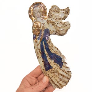 May include: A ceramic angel ornament featuring a glossy cream-colored glaze with brown speckled accents. The figure has a gold-painted halo, a blue robe, and outstretched wings. The surface has a textured, hand-crafted appearance with visible glaze variations.