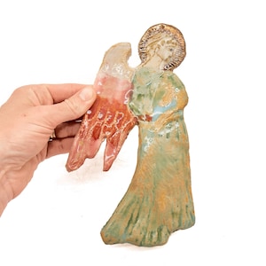 Può includere: Una statuetta di angelo in ceramica scolpita a mano con finitura lucida. L'angelo presenta una veste verde testurizzata, un'ala prominente con smalto rosso-arancio e un'aureola dorata attorno alla testa. La superficie mostra la texture dell'argilla.