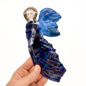 May include: A decorative ceramic angel figurine featuring a glossy finish with deep blue and iridescent purple tones. The figure has a painted face with a metallic gold halo, large textured blue wings, and a flowing gown with a dark, reflective glaze.
