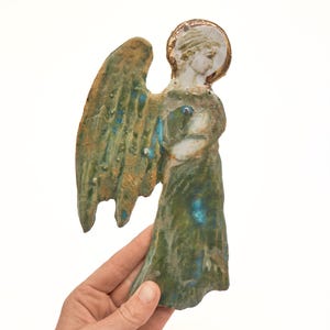 Op de afbeelding: Een plat keramieken engelfiguur met een textuurrijke groen-bruine glazuurlaag en blauwe accenten. De engel heeft een wit gezicht, een gouden halo en grote vleugels, afgebeeld in zijaanzicht met de handen gevouwen bij de borst.