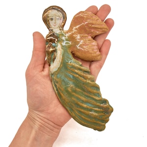 May include: A hand-sculpted ceramic angel figurine featuring a glossy finish with a green and gold flowing gown, a textured tan wing, and a painted face with a golden halo. The piece displays a mix of earthy tones and metallic gold accents.