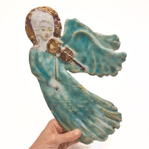 Può includere: Una placca da parete in ceramica a forma di angelo che suona il violino. La figura presenta un volto bianco, un'aureola dorata e una veste fluida smaltata in tonalità blu e verde acqua. L'angelo ha grandi ali spiegate e un lungo abito plissettato.