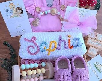 May include: A pink baby gift box with a pink baby outfit, a white blanket with the name "Sophia" embroidered in colorful letters, a wooden toy with colorful beads, a pair of pink baby shoes, and a wooden block with the text "3 Months".