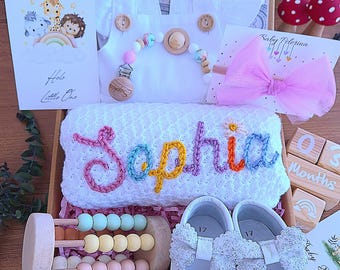 May include: A baby gift set in a wooden box includes a white romper, a personalized blanket embroidered with "Sophia", a pair of white shoes, a pacifier clip, a hair bow, a card that says "Hello Little One", and a wooden abacus. The set is decorated with pastel colors.