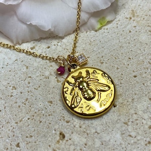 Può includere: Collana in oro con ciondolo a forma di ape e piccole pietre rosa e trasparenti. Il ciondolo a forma di ape è rotondo e presenta un design dettagliato.