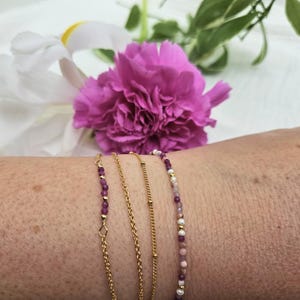 May include: Four gold bracelets stacked on a wrist. The top bracelet is a delicate chain with small pink and white beads. The second bracelet is a thicker chain with small gold beads. The third bracelet is a delicate chain with small purple beads. The bottom bracelet is a delicate chain with small purple beads.