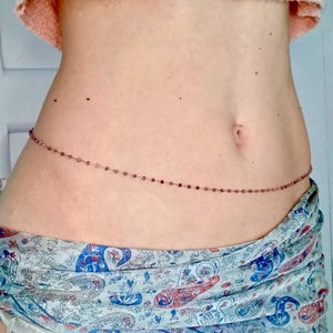 Może przedstawiać: Delikatny łańcuszek na talię z małymi, czerwonobrązowymi koralikami. Koraliki są nawleczone, tworząc delikatny dodatek. Łańcuszek jest udrapowany wokół talii, co sugeruje, że jest noszony jako biżuteria na ciało. Koraliki są równomiernie rozmieszczone.