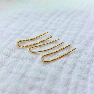 Może przedstawiać: Trzy złote kolczyki z ozdobnym splotem. Kolczyki mają różne długości i zakrzywiony kształt.