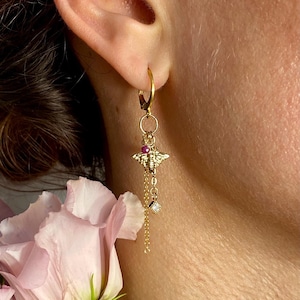 May include: Gold bee charm earrings with a dangling chain and a small clear crystal. The earrings are hanging from a person's ear.