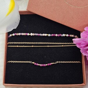 May include: A set of three delicate gold chain bracelets with pink and purple gemstone beads. The bracelets are displayed in a brown gift box.