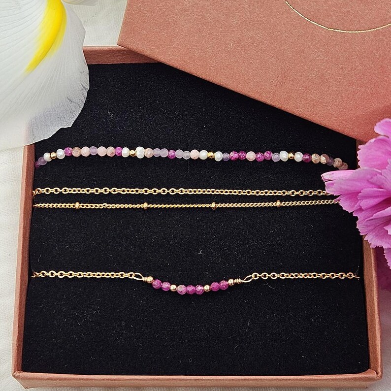 May include: A set of three delicate gold chain bracelets with pink and purple gemstone beads. The bracelets are displayed in a brown gift box.