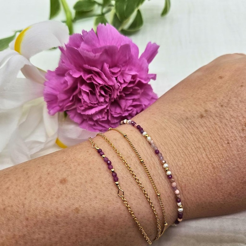May include: Four gold bracelets on a wrist. The bracelets are layered, with two delicate chain bracelets and two beaded bracelets. One beaded bracelet has pink and white beads, and the other has purple beads.