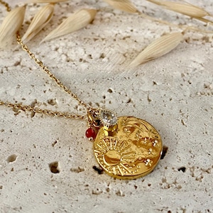 Op de afbeelding: Gouden ketting met een ronde hanger met een zon- en maandesign met kleine sterren en een heldere edelsteen. De hanger is bevestigd aan een delicate gouden ketting.