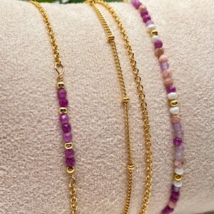 May include: Three gold chain bracelets with purple and pink gemstone beads. The bracelets are layered on a pink fabric background.