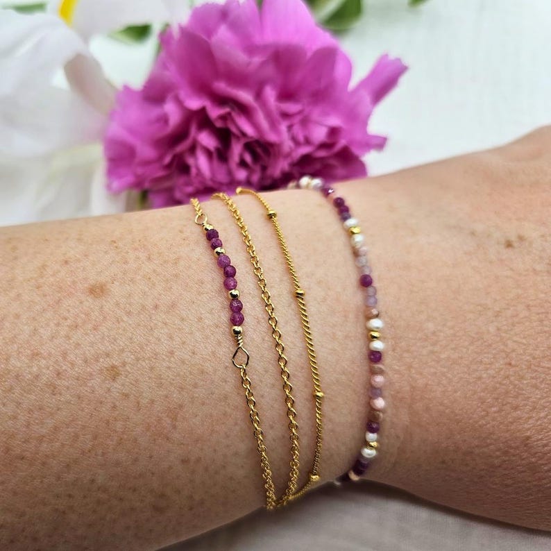 May include: Four gold bracelets stacked on a wrist. The top bracelet has pink and white beads. The second bracelet has small purple beads. The third bracelet is a gold chain with small gold balls. The bottom bracelet is a gold chain with a twisted design.