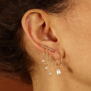 Op de afbeelding: Een gouden oormanchet met een rij kleine parels die eraan hangen. De manchet wordt op het bovenste deel van het oor gedragen, en een parel oorbel wordt op de onderste oorlel gedragen.