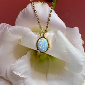 Peut inclure: Un collier délicat en chaîne dorée avec un pendentif en opale blanche. Le pendentif est serti dans une bélière dorée et a une forme légèrement irrégulière. Le collier est photographié sur une fleur blanche.