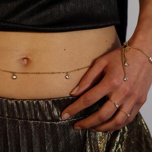 Könnte beinhalten: Eine goldene Kette mit kleinen, klaren Edelsteinen, die daran hängen. Die Kette wird um die Taille getragen und hat einen baumelnden Anhänger an der Seite.