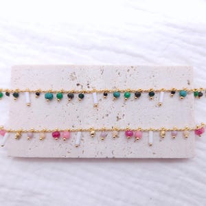 May include: Two gold chain bracelets with colorful beads and white beads hanging from the chain. The top bracelet has green and black beads, while the bottom bracelet has pink and white beads.