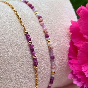 May include: Two delicate bracelets with gold chain accents. One bracelet features pink, purple, and white beads, while the other features purple beads.