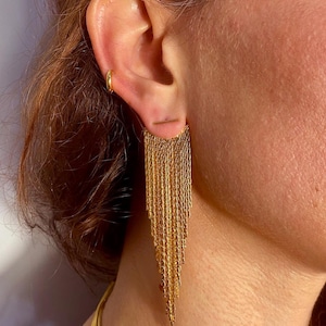 Può includere: Orecchini pendenti dorati con un lungo design a catena. Gli orecchini sono realizzati con più catene sottili che pendono da una piccola barra.