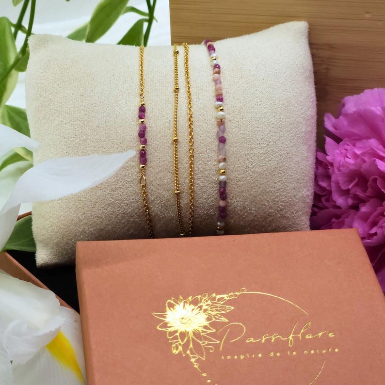 May include: Four gold chain bracelets with pink and white beads. The bracelets are displayed on a pink cushion and a brown box with the text "Parrflare inspired de la nature".