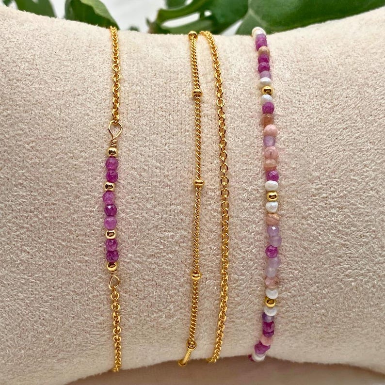 May include: Four gold bracelets with pink and purple beads. The bracelets are layered on a pink fabric background.