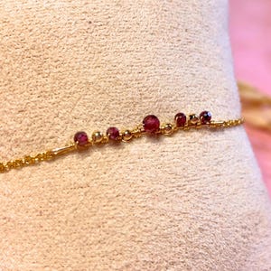 Peut inclure: Un bracelet en or délicat orné de petites pierres précieuses rondes et rouge foncé. De minuscules perles d'or et une fine chaîne en or relient les pierres précieuses. Le bracelet est présenté sur une surface de couleur neutre.
