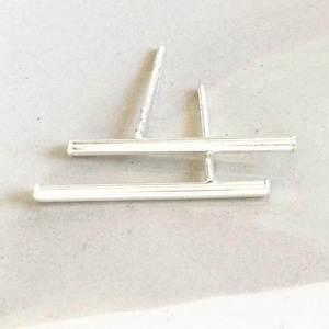 Puede incluir: Un par de pendientes de barra plateados con un diseño simple y minimalista. Los pendientes están hechos de dos barras finas y rectas que se cruzan en ángulo recto.