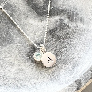 May include: A silver necklace with a small round pendant featuring the letter 'A' and a light blue gemstone.