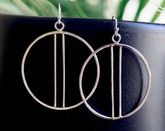 Puede incluir: Pendientes de aro plateados con un diseño minimalista. Cada pendiente presenta un círculo grande con dos barras verticales en el interior. Los pendientes se muestran sobre un fondo negro con follaje verde.