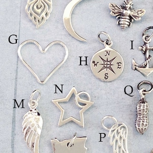May include: A collection of 16 silver charms, including a crescent moon, a heart, a star, a compass, a bee, a feather, a peanut, a wing, and an anchor. The charms are arranged on a light gray background.