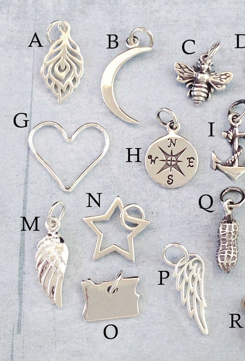 May include: A collection of 16 silver charms, including a crescent moon, a heart, a star, a compass, a bee, a feather, a peanut, a wing, and an anchor. The charms are arranged on a light gray background.