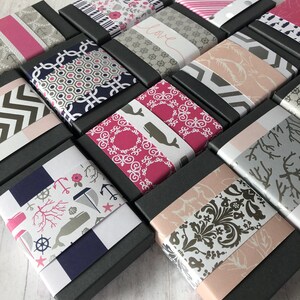 May include: A collection of small, black gift boxes with various patterned paper lids. The patterns include anchors, whales, flowers, and geometric designs. The boxes are arranged in a staggered pattern on a white wooden surface.
