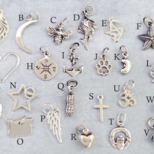 May include: A collection of 26 silver charms, each featuring a different design. The charms include a feather, crescent moon, bee, fish, dolphin, starfish, heart, compass, anchor, paw print, crescent moon, lotus flower, angel wing, star, peanut, cross, Om symbol, pearl, and a bell.