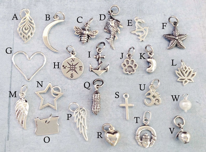 May include: A collection of 26 silver charms, each featuring a different design. The charms include a feather, crescent moon, bee, fish, dolphin, starfish, heart, compass, anchor, paw print, crescent moon, lotus flower, angel wing, star, peanut, cross, Om symbol, pearl, and a bell.
