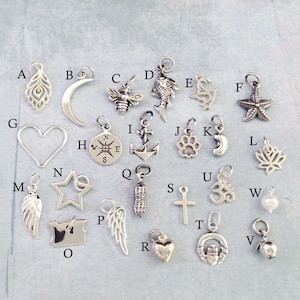 May include: A collection of 26 silver charm pendants, each featuring a different design, including a heart, a star, a feather, a compass, a cross, a paw print, a fish, a bee, a crescent moon, a starfish, a peanut, a leaf, a wing, a lighthouse, a bell, an anchor, an Om symbol, and a state of Oregon.