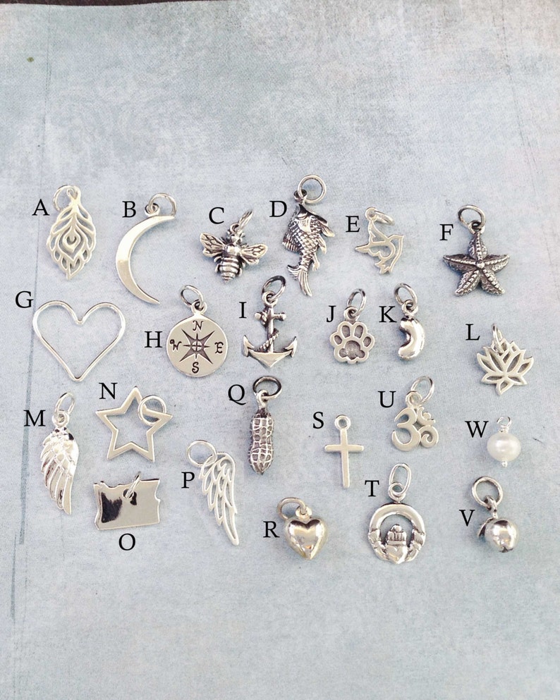 May include: A collection of 26 silver charm pendants, each featuring a different design, including a heart, a star, a feather, a compass, a cross, a paw print, a fish, a bee, a crescent moon, a starfish, a peanut, a leaf, a wing, a lighthouse, a bell, an anchor, an Om symbol, and a state of Oregon.