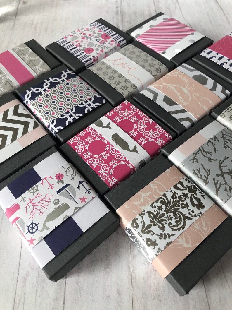 May include: A collection of small, black gift boxes with various patterned paper lids. The patterns include anchors, whales, flowers, and geometric designs. The boxes are arranged in a staggered pattern on a white wooden surface.