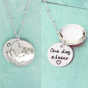 Peut inclure: Deux colliers en argent avec des pendentifs ronds. Un pendentif est martelé et l'autre porte le texte "One day closer" avec un petit cœur.