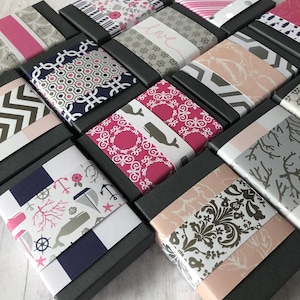 May include: A collection of small, black gift boxes with various patterned paper lids. The patterns include anchors, whales, flowers, and geometric designs. The boxes are arranged in a staggered pattern on a white wooden surface.