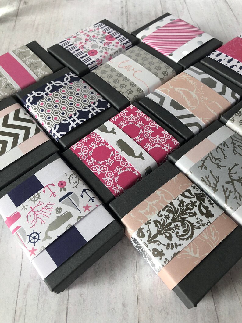 May include: A collection of small, black gift boxes with various patterned paper lids. The patterns include anchors, whales, flowers, and geometric designs. The boxes are arranged in a staggered pattern on a white wooden surface.